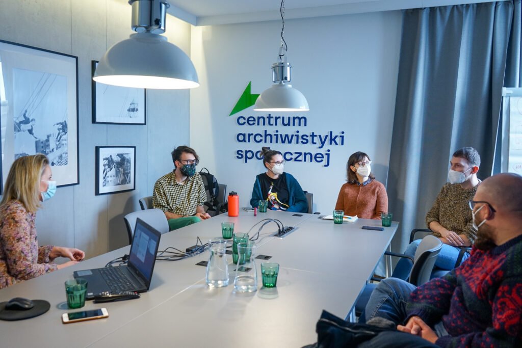 Sześć osób siedzi wokół stołu konferencyjnego. Na twarzach mają maseczki, słuchają jednego z mężczyzn. Za nimi ściany z oprawionymi zdjęciami oraz logo Centrum Archiwistyki Społecznej. 