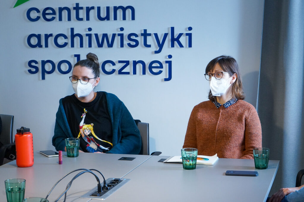 Dwie gościnie z Czeskiej Akademii Nauk siedzą w maseczkach przy stole. Za nimi logo CAS. 
