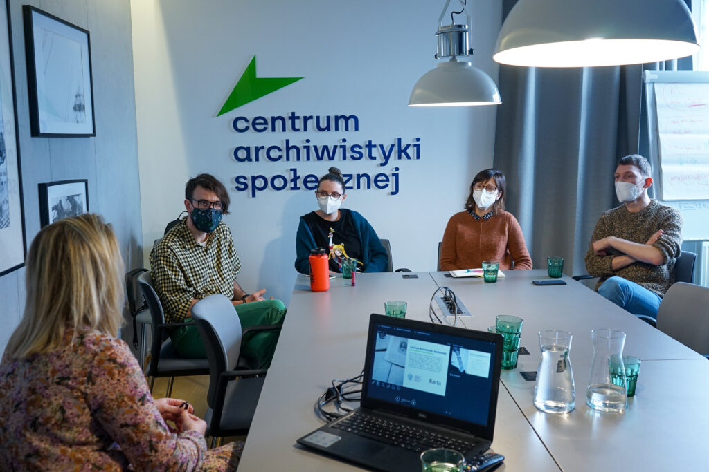 Trzy kobiety i dwóch mężczyzn przy stole. Na stole leży też laptop, na ekranie prezentacja. Za nimi ściany z oprawionymi zdjęciami, flipchart z notatkami oraz logo Centrum Archiwistyki Społecznej.